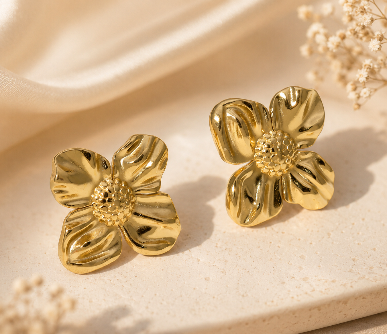 Gold flower-shaped earrings on a beige surface with dried flowers.