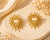 Gold sunburst earrings with heart design on a beige stone surface.